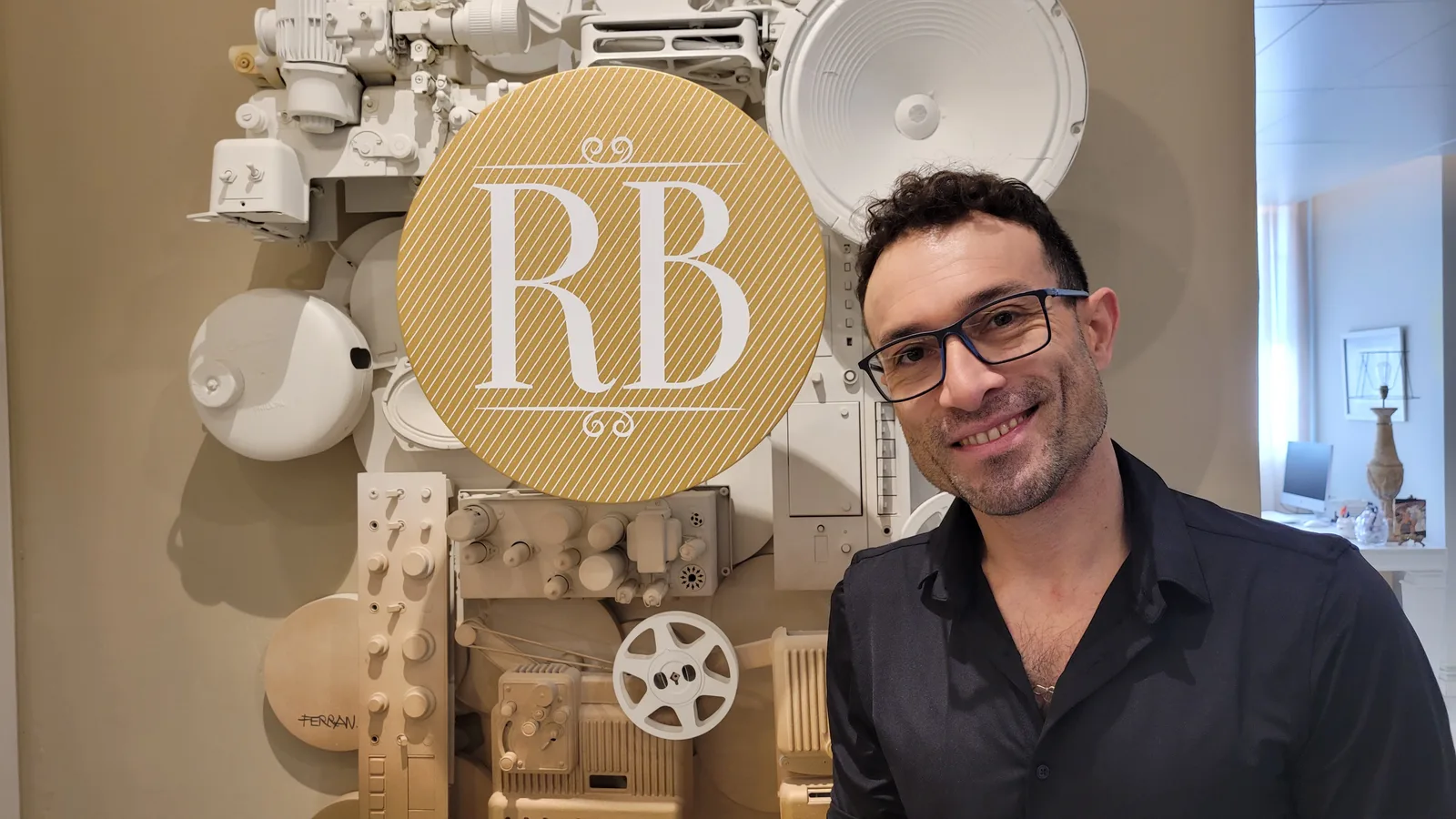 Ricardo Buriticá sonriendo junto al mural del estudio, con el sello RB en dorado